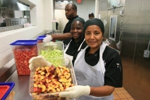 School cafeteria workers holding large tubs of fresh cut fruit.