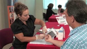 Woman in black performing a finger stick for a cholesterol test on a man in a blue plaid shirt.