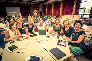 Group of women attending a conferenece gathered at a large table with presentation style seating behind them.