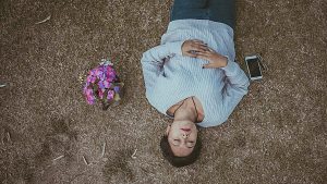 Woman in a light grey shirt laying outside meditating with eyes closed next to her phone and purple flowers.