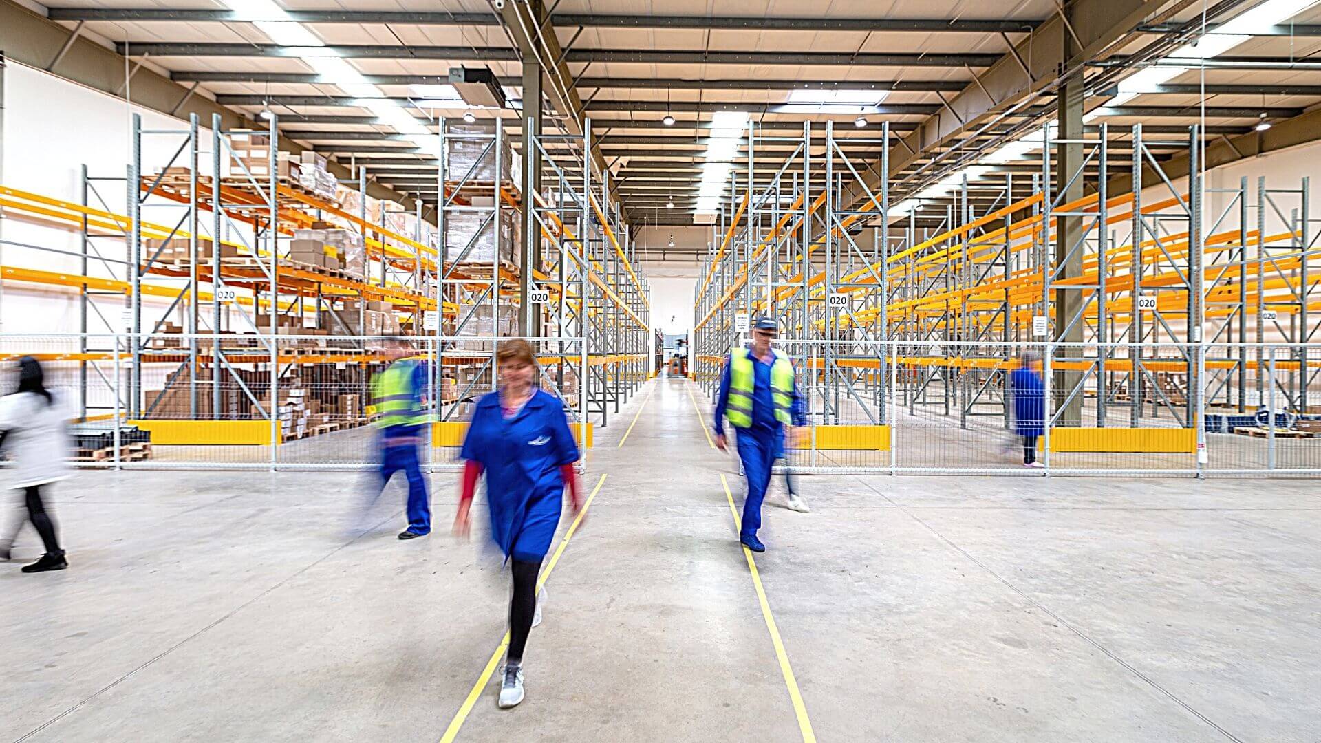 Multiple warehouse workers in blue with yellow caution vests walking through an empty warehouse. wellness programs improve employee retention
