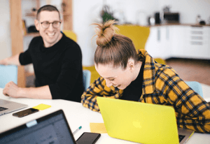 Two people sitting at a desk laughing and working on laptops.