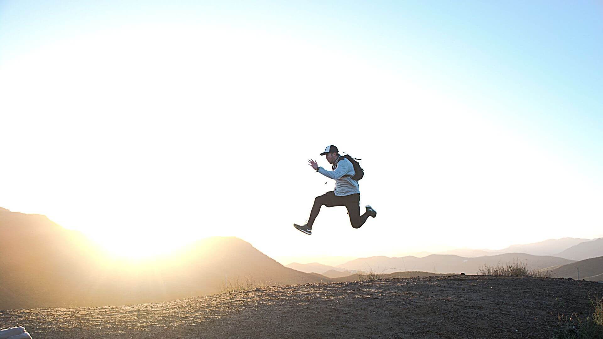 Man in a hat on a trail jumping high into the air as if he is floating in front of a large mountain range. 10 Effective Ways to Motivate Employees in the Workplace