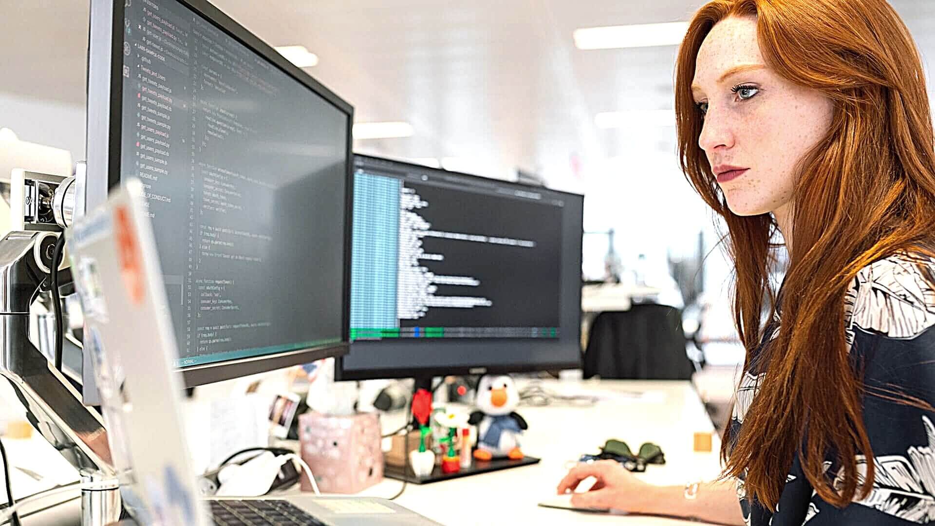 Side profile of a woman with red hair working at a desk with three large screens.