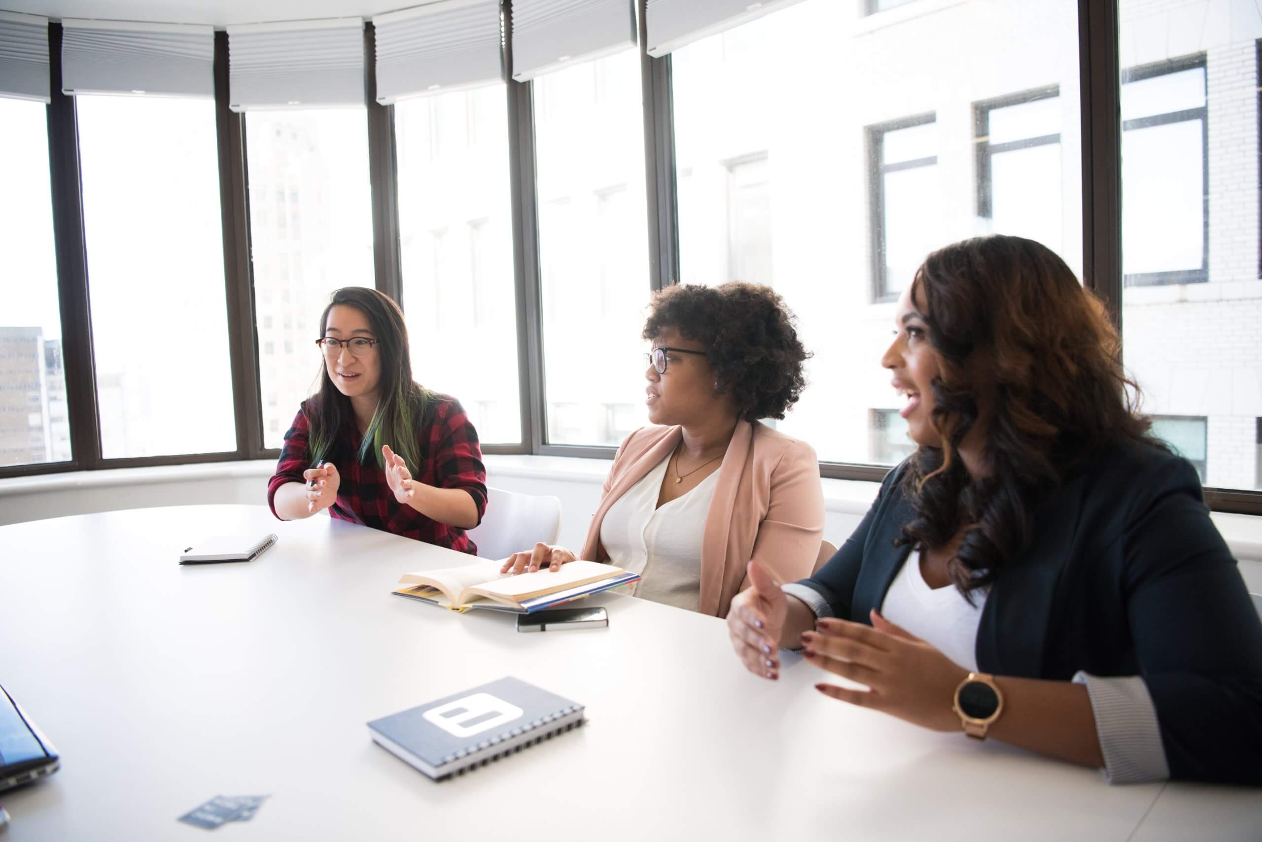 Three corporate women conversing at a large table in an office surrounded by windows.