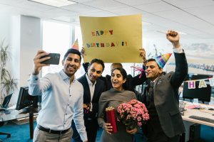 Employees group celebrating a birthday. Some wearing party hats while others hold a sign for a picture.