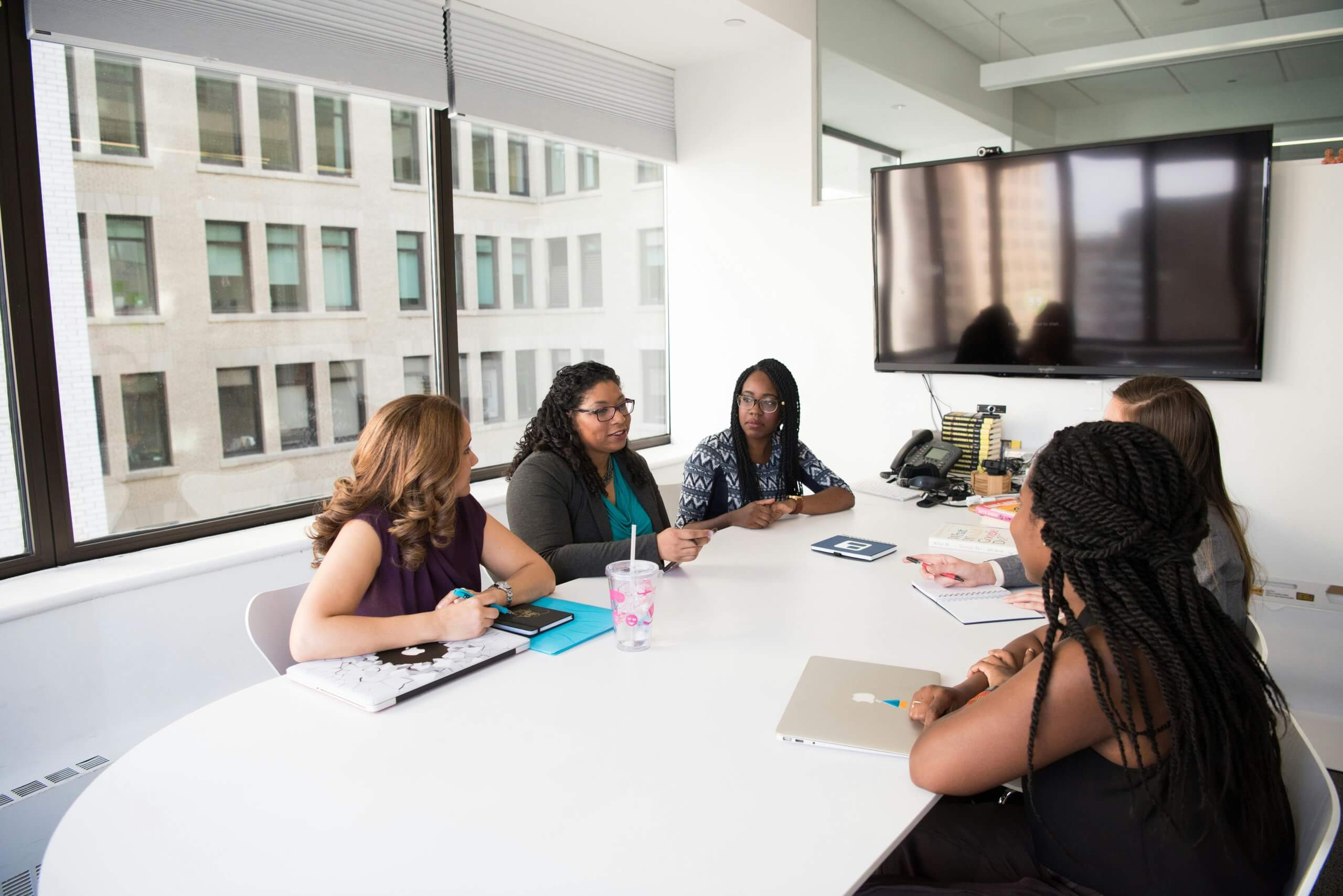 Group of 5 women sitting at a conference table discussing DEI activities in front of a large TV and window.