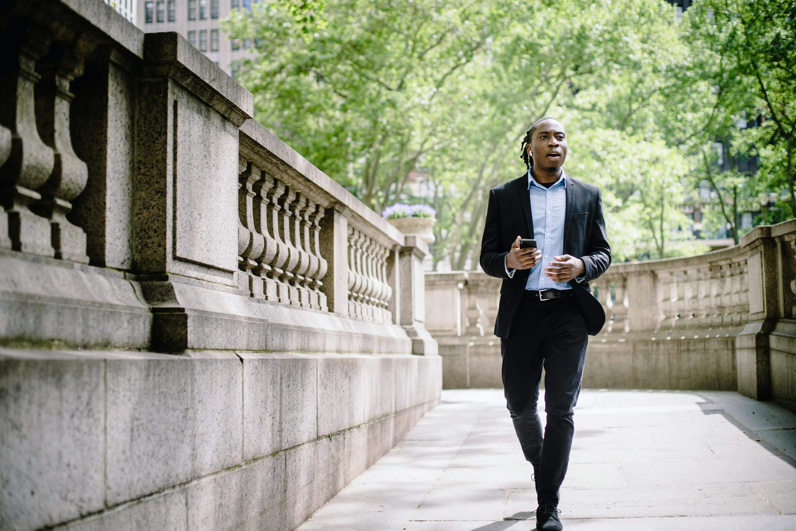 African American man walking through a city park taking a meeting on his phone.